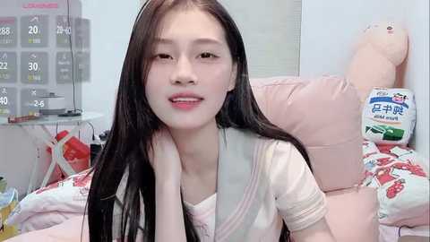 Video of a young East Asian woman with long black hair, smiling, wearing a white shirt, lying on a pink bed with cartoon pillows, in a cozy, cluttered bedroom.