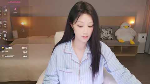 Video of a young East Asian woman with long black hair, wearing a light blue striped pajama top, sitting on a beige bed in a modern, minimalist bedroom.
