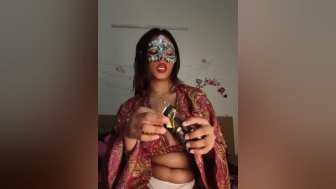 Video of a woman with medium skin tone and long dark hair, wearing a silver masquerade mask, red lipstick, and a vibrant, floral-patterned kimono. She holds a black pistol with both hands.