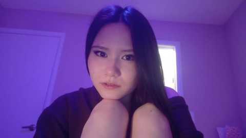 Video of an East Asian woman with long black hair, light skin, and dark eyes, wearing a dark top, sitting on a bed, indoors with purple lighting, closed door and window in the background.