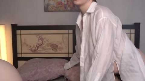Video of a young, light-skinned person in a white, open-fronted nightgown kneeling on a bed with pink pillows. Background features a wooden headboard with Asian-style artwork.