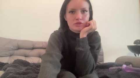 A video of a woman with long black hair, resting her chin on her hand, wearing a dark sweater, seated on a gray, tufted sofa.