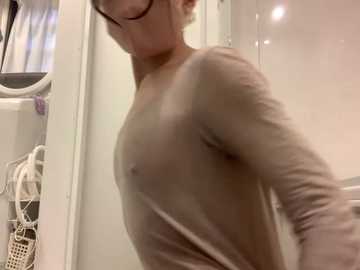 A video of a slender, fair-skinned woman with short hair, wearing a sheer, long-sleeved top, standing in a white-tiled bathroom with a washing machine in the background.