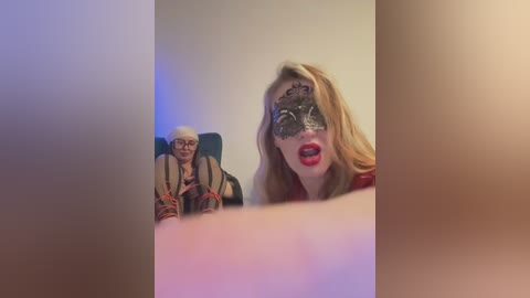 Media: Video of a woman with blonde hair, wearing a black lace mask, and red lipstick, sitting in a chair. An elderly man with glasses and a striped shirt sits behind her.