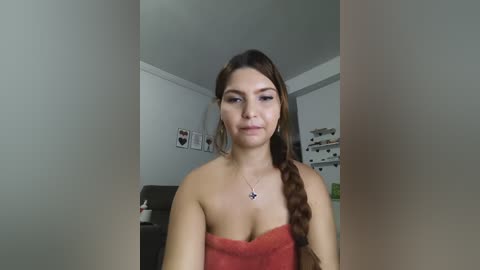 A video of a young woman with long brown hair in a braid, wearing a red towel and a necklace, standing in a modern living room with white walls, a gray couch, and a shelf of books.