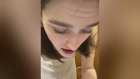 A video of a young woman with straight, dark hair, wearing a light grey, ribbed sweater, looking downward with an open-mouth expression, framed by a blurred, warm-toned background.