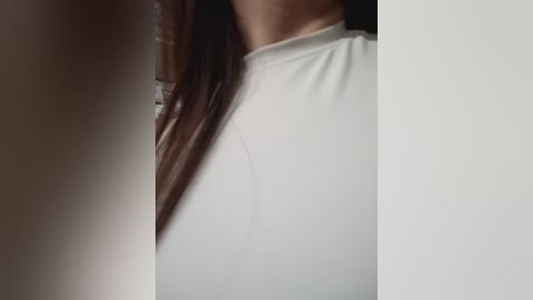 A close-up video of a woman's neck and shoulders, wearing a white, high-necked top. Her long, straight brown hair cascades over her left shoulder. The background is out of focus.