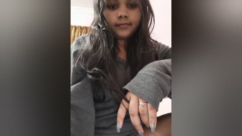 Video of a young Black woman with long, wavy black hair, wearing a gray sweater, with a silver ring on her left hand, seated indoors against a blurred background.