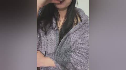 Video of a person with long black hair, wearing a grey, textured bathrobe, partially covering their face with one hand. Background is blurred, featuring a gradient from dark to light grey.