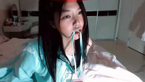 Video of a young Asian woman with long black hair, wearing a light blue dress, lying on a bed with white sheets, holding a blue tube to her mouth, in a modern bedroom with a white door and window.