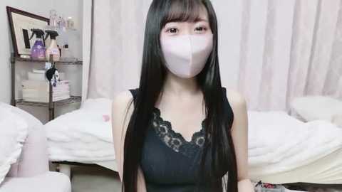 Video of an East Asian woman with long black hair and a pale complexion, wearing a black lace-trimmed top, face mask, and seated in a white-tiled room with unmade beds.