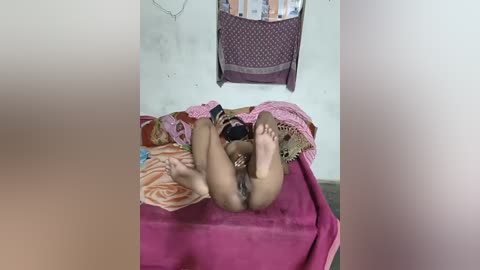 Video of a naked woman with medium skin tone, lying on a bed in a sparsely decorated room. She's holding a black smartphone and has her legs bent, exposing her genitals.