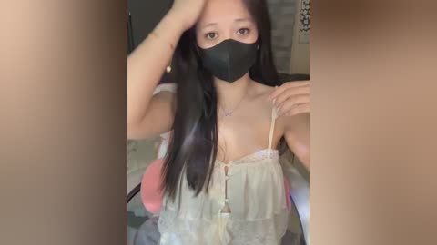Video of an East Asian woman with long black hair, wearing a black face mask, beige lace camisole, and sitting in a chair.