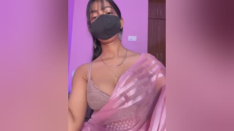 Video of a South Asian woman with medium skin tone, wearing a black face mask, light pink saree, and a beige lace bra, standing in a room with purple walls and a wooden door.