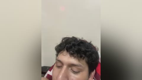 A video shows a young man with curly dark hair, wearing a red and white striped shirt, lying on a bed with his eyes closed. The background is a plain, light-colored wall.