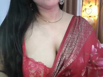 Video of a light-skinned woman with long, dark hair, wearing a red sari with intricate floral embroidery and a lacy red bralette, adorned with gold jewelry, in a softly lit room with warm tones.