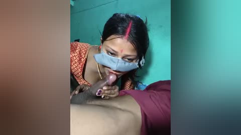 A video shows a South Asian woman in a mask performing oral sex on a man lying on a bed, with teal walls in the background.
