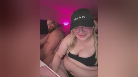 Video of a plus-sized, blonde woman in glasses and a black cap, smiling, with a muscular, bearded man behind her, both nude, in a dimly lit room with pink lighting.