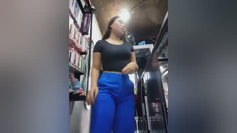 Video of a woman in a cluttered, dimly lit storage room, wearing a black top and bright blue pants. Shelves lined with boxes and a fridge in the background.