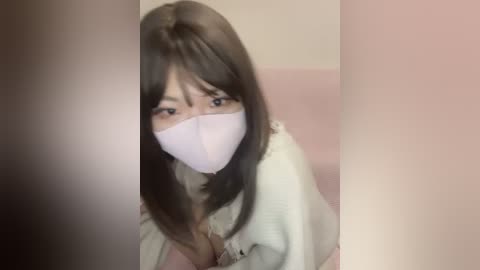 A video of a young woman with long, straight dark brown hair and a white surgical mask, wearing a white robe, seated in a beige armchair. The background is out of focus, featuring a soft pink wall.