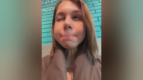 Media: Video of a young woman with light skin, straight brown hair, and a neutral expression, wearing a light brown jacket. Background features a teal brick wall with \"PIZZA\" written in white.