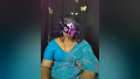Media: Video of a plus-size woman in a blue saree, wearing a purple and black Venetian mask, with butterflies on a dark curtain backdrop.