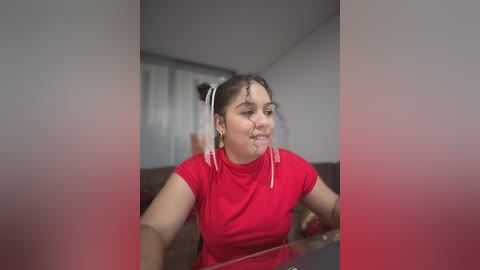Video of a young woman with dark hair in a red t-shirt, messy makeup, and a blurred background in a dimly lit room.