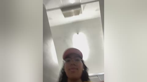 A blurry photo of a young woman with long black hair and glasses, wearing a pink hat, standing in a narrow, brightly lit corridor with white walls and fluorescent lights.