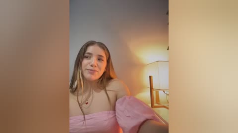 A video of a young Caucasian woman with long, straight brown hair, wearing a pink off-shoulder top, smiling. She's framed by a light beige background, with a warm-toned lamp and a white lampshade in the background.