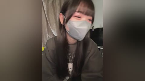 Video of a young person with long dark hair, wearing a gray face mask and a dark jacket, looking down with a solemn expression, indoors.