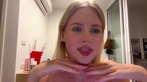 A video of a blonde woman with fair skin, blue eyes, and parted lips, leaning forward with hands on her chest, in a modern, minimalistic room with white walls, a red shelf, and a partially visible door.