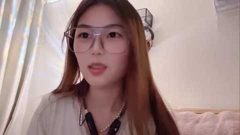 Video of an Asian woman with long brown hair, wearing clear glasses, a white blouse, and a pearl necklace, in a bathroom with a shower curtain, towel, and a black wire rack.