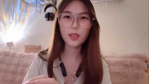 Video of an East Asian woman with long brown hair, wearing glasses and a white top, with a microphone and headset. She sits on a beige couch in a studio with soft lighting and a box in the background.