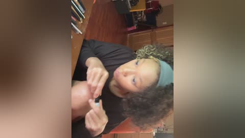 A video of a young woman with curly hair, wearing a headband and a black top, holding a phone, in a cluttered room with wooden furniture and books.