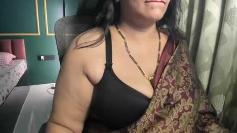 Video of a woman with medium skin tone and black hair, wearing a black bra and gold necklace, seated in a modern bedroom with green walls, a bed, and patterned curtains.