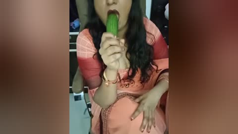 Video of a woman with long black hair, wearing a pink, embroidered dress, eating a green cucumber with her mouth open.