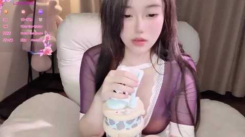 Media: Video of a young Asian woman with fair skin, long black hair, wearing a sheer purple robe, holding a blue and white macaron, seated on a white chair in a dimly lit room.
