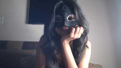 Video of a woman with long black hair, wearing a black mask with silver accents, resting her chin on her hand, smiling. Background shows a beige wall, a flat-screen TV, and a patterned couch.