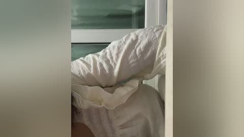 Video of a messy white bed with wrinkled sheets and a pillow, partially visible through a frosted glass window, creating a soft, muted ambiance.