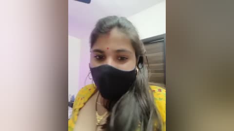 Video of a young woman with medium brown skin, wearing a yellow floral blouse, black mask, and red dot on her forehead, taken indoors.