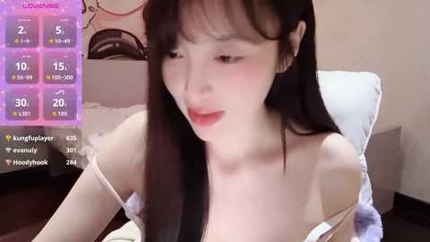 A video of a young East Asian woman with long black hair, wearing a white tank top, reading a book in a cozy bedroom.