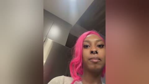 Video of a young Black woman with bright pink hair, silver septum piercing, and a neutral expression, standing in a modern, minimalist hallway with metallic walls and a soft, blurred background.