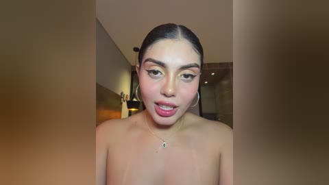 Video of a young woman with medium skin tone, dark hair pulled back, wearing hoop earrings and a necklace, posing topless indoors, smiling with her tongue out.