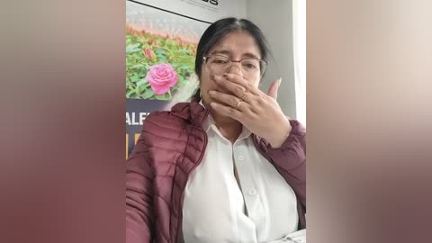 A video of a South Asian woman with glasses, maroon puffer jacket, white blouse, and hand over mouth, standing in a room with a flowered poster in the background.