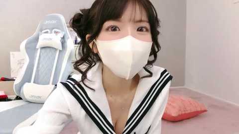 Video of an East Asian woman with dark hair, wearing a white sailor uniform and face mask, sitting in a gaming chair with a pink pillow on a light carpeted floor.
