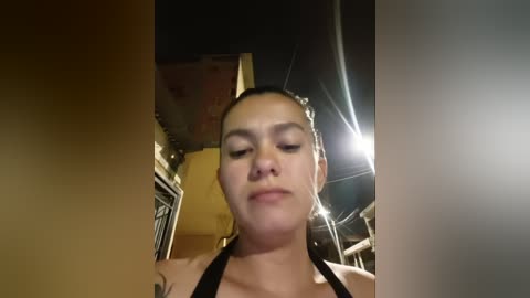 Media: Video of a young woman with light skin, dark hair in a bun, wearing a black halter top, indoors with dim lighting, blurry background.