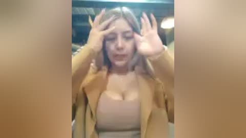 A video shows a woman with fair skin, long blonde hair, and a tan coat, holding her face with both hands, looking distressed. The background is blurred with a warm, indoor setting.