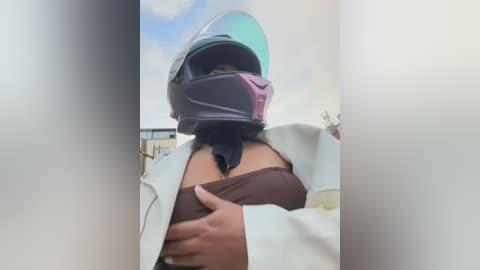 A video of a person wearing a black motorcycle helmet with a purple visor, exposing their bare chest. They have medium-brown skin, and their left hand is touching their exposed breast. The background shows a partly cloudy sky and blurred outdoor scenery.