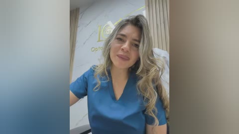 Video of a light-skinned woman with wavy blonde hair, wearing a blue V-neck top, standing in a bright, modern dental office with marble walls and a golden logo.
