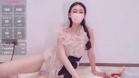 Video of an Asian woman in a ruffled, sheer pink dress, wearing a face mask, lying on a floor with a white background.